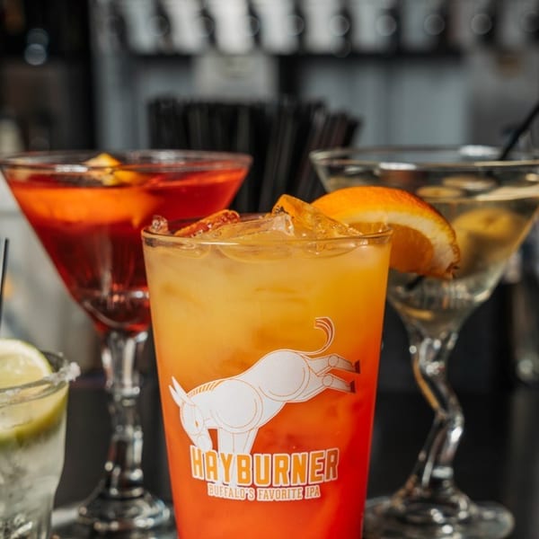 Four colorful cocktails, including a Hayburner IPA glass, on a dark bar surface.